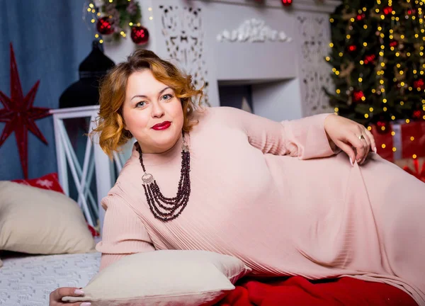 Uzun pembe elbiseli güzel bir tombul kız yeni yıl süslemeleri ile dekore edilmiş bir odada bir yatakta yatıyor.