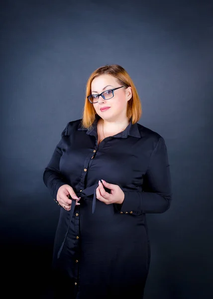 Karanlık bir arka plan üzerinde siyah elbiseli güzel tombul kız. Kendine güvenen artı boyutu kadın gözlük ve çerçeve içinde poz siyah elbise