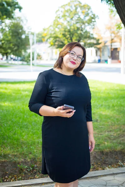 Siyah elbiseli Buxom kız bir parkta elinde bir telefon ile duruyor