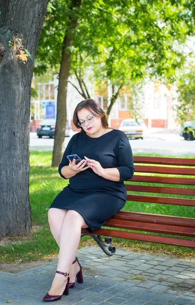 Siyah sıkı elbise güzel tombul kız elinde bir telefon ile parkta bir bankta oturur