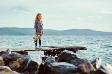 Bir oyuncakla üzgün bir çocuk deniz kenarındaki iskelede duruyor ve su sıçramalarına görünüyor