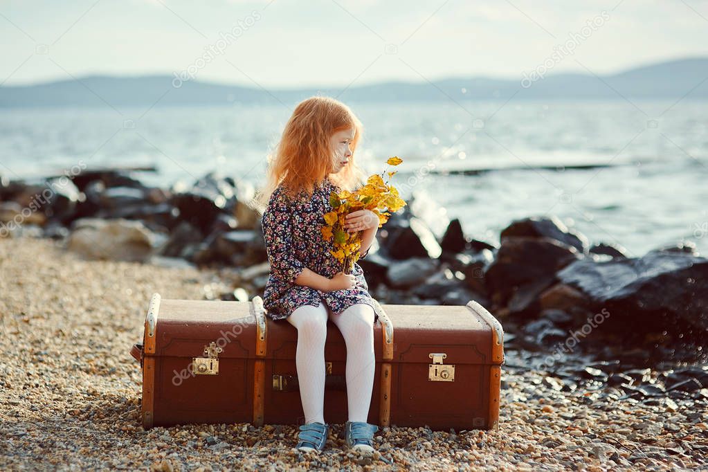A little sad girl with a toy sitting on a big suitcase near the sea