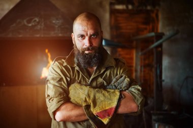 Bir vahşi, kendinden emin demirci, sakallı bir adam portresi. Atölye çalışmasında, kollarını göğsüne üzerinde katlanır ayakta.