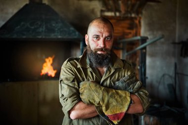Bir vahşi, kendinden emin demirci, sakallı bir adam portresi. Atölye çalışmasında, kollarını göğsüne üzerinde katlanır ayakta.