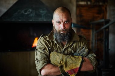 Bir vahşi, kendinden emin demirci, sakallı bir adam portresi. Atölye çalışmasında, kollarını göğsüne üzerinde katlanır ayakta.