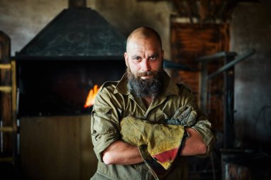 Bir vahşi, kendinden emin demirci, sakallı bir adam portresi. Atölye çalışmasında, kollarını göğsüne üzerinde katlanır ayakta.