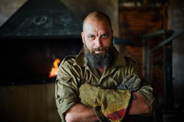 Bir vahşi, kendinden emin demirci, sakallı bir adam portresi. Atölye çalışmasında, kollarını göğsüne üzerinde katlanır ayakta.