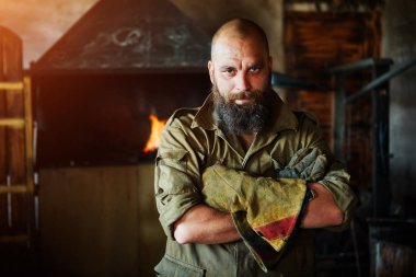 Bir vahşi, kendinden emin demirci, sakallı bir adam portresi. Atölye çalışmasında, kollarını göğsüne üzerinde katlanır ayakta.