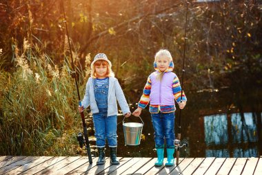 İki kız köprünün üzerinde durmak ve onların elleri balıkçılık çubuklar ve bir kova balık için basılı tutun.