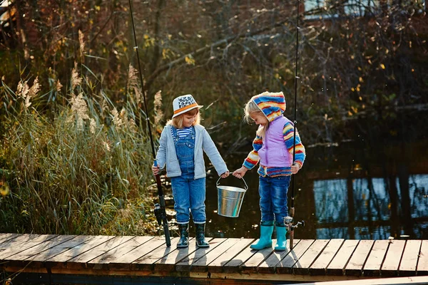Дві маленькі дівчинки з рибальськими паличками стоять на дерев'яному понтоні і дивляться на рибу, спійману у відро — стокове фото