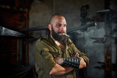 Acımasız sakallı demirci işçi zor işten sonra sigara içiyor. Adam yorgun ama gülümseyen, yüzünü çamur ve ter kaplıdır