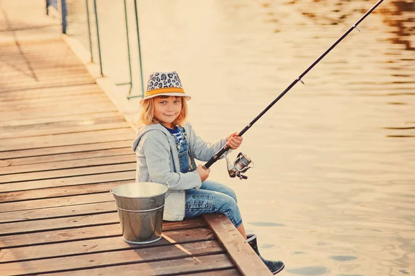 Schattig kind meisje vissen vanaf houten pier op een meer. Familie vrijetijdsbesteding in de zomer zonnige dag. Meisje vermaakt zich bij een rivier op een mooie zomeravond. Actieve familietijd over de natuur — Stockfoto