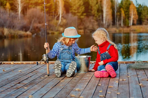 Dos hermanitas o amigos se sientan con cañas de pescar en un muelle de madera. Cogieron un pez y lo pusieron en un cubo. Ellos están contentos con su captura y discutirlo . —  Fotos de Stock