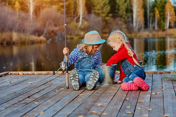 Dos hermanitas o amigos se sientan con cañas de pescar en un muelle de madera. Cogieron un pez y lo pusieron en un cubo. Ellos están contentos con su captura y discutirlo . — Foto de Stock