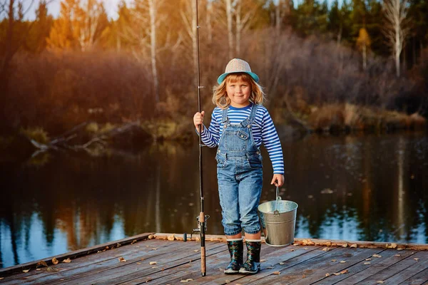 Niña pescadora con una caña de pescar y un cubo de pescado en un muelle de madera —  Fotos de Stock