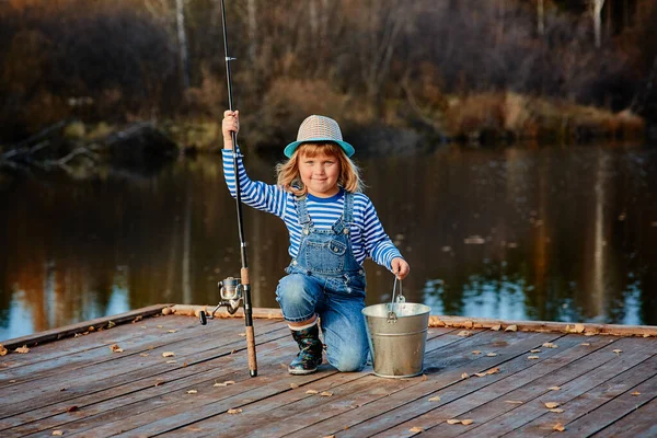 Niña pescadora con una caña de pescar y un cubo de pescado en un muelle de madera — Foto de Stock