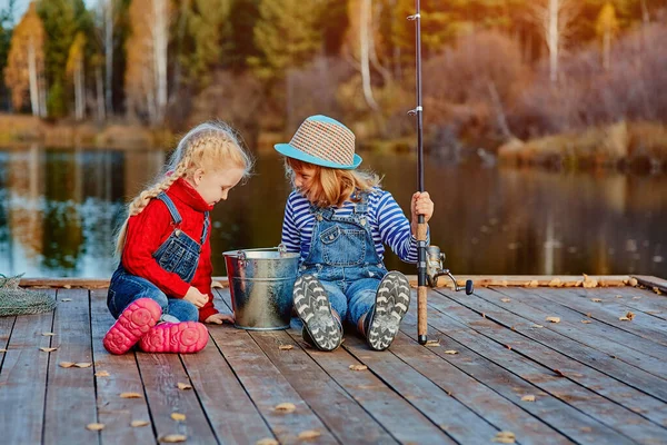 Δύο μικρές αδελφές ή φίλοι κάθονται με καλάμια ψαρέματος σε μια ξύλινη προβλήτα. Έπιασαν ένα ψάρι και το έβαλαν σε έναν κουβά. Είναι ευχαριστημένοι με την ψαριά τους και το συζητούν.. — Φωτογραφία Αρχείου