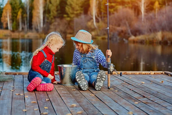 Dos hermanitas o amigos se sientan con cañas de pescar en un muelle de madera. Cogieron un pez y lo pusieron en un cubo. Ellos están contentos con su captura y discutirlo . — Foto de Stock