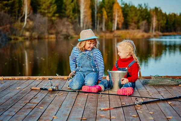 Dos hermanitas o amigos se sientan con cañas de pescar en un muelle de madera. Cogieron un pez y lo pusieron en un cubo. Ellos están contentos con su captura y discutirlo . — Foto de Stock