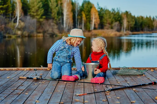 Dos hermanitas o amigos se sientan con cañas de pescar en un muelle de madera. Cogieron un pez y lo pusieron en un cubo. Ellos están contentos con su captura y discutirlo . — Foto de Stock