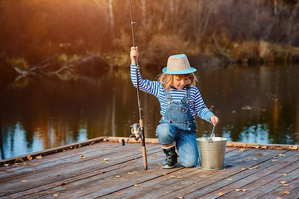 Una niña con una caña de pescar mira la captura de peces en un cubo —  Fotos de Stock