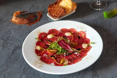 Ahşap zemin üzerinde beyaz bir tabakta et carpaccio, lezzetli bir ekmek ve bir bardak şarap ile.