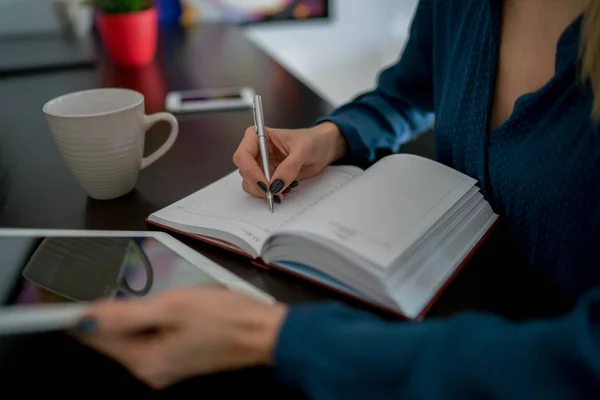 Female author of journalism field writing new article down lined paper pages of notebook. Close up view of women\'s hands holding white touch pad with reflection on screen. Use of modern technologies