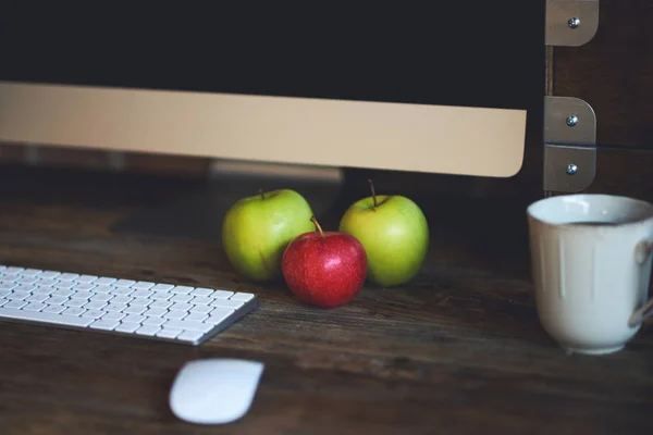 Comfortable workplace with big computer screen, mouse and keyboard. Wooden table with pencils for drawing and colorful apples.  Desktop with  cup of coffee and fresh apples for healthy breakfast.