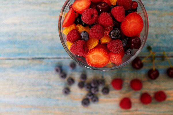 Top view on a vintage wooden desk with peeling paint, there is a deep glass bowl with fresh chopped salad of ripe berries