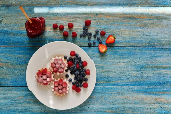 Cook cakes served on a white plate, tartlets with raspberry jam and fresh raspberries sprinkled icing sugar