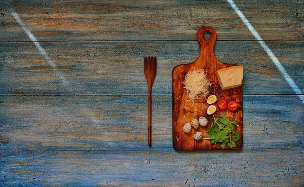 On a wooden shabby retro table is a wooden board for cutting vegetables on it is a piece of Parmesan cheese, quail eggs and cherry tomatoes, near a large wooden fork