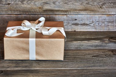 El yapımı hediye kutusu gri ahşap rustik arka plan üzerinde beyaz saten kurdele ile Kraft kağıt sarılı. Kurdele ile yılbaşı, Sevgililer günü, Doğum günü veya yeni yıl için herhangi bir tatil kavramı sunar. 
