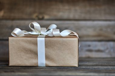 El yapımı hediye kutusu gri ahşap rustik arka plan üzerinde beyaz saten kurdele ile Kraft kağıt sarılı. Kurdele ile yılbaşı, Sevgililer günü, Doğum günü veya yeni yıl için herhangi bir tatil kavramı sunar. 