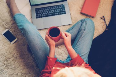 Laptop ve kahve Kupası ahşap bir katta oturan kızın elinde. Dizüstü bilgisayar, tablet ve kahve Kupası'nda rahat ev yazarak eller kadın. Üstten görünüm genç işkadını parlak modern iş istasyonunda oturan.