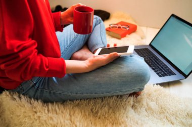 Laptop ve kahve Kupası ahşap bir katta oturan kızın elinde. Dizüstü bilgisayar, tablet ve kahve Kupası'nda rahat ev yazarak eller kadın. Yan görünümü genç iş kadını evde katta oturan.