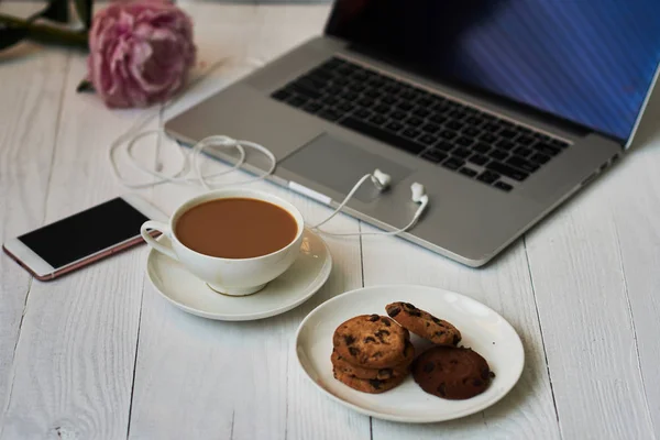 Hot cup of fresh coffee and laptop on the work desk. Cosy home and work concept - vintage wooden table with laptop computer, coffee cup and biscuit. Women\'s elegant desktop. Selective focus.