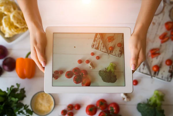 View of female hands holding white digital tablet with colorful photo on the screen of healthy salad recipe with vegetables. Selective focus on touch pad on which female food blogger taking picture.