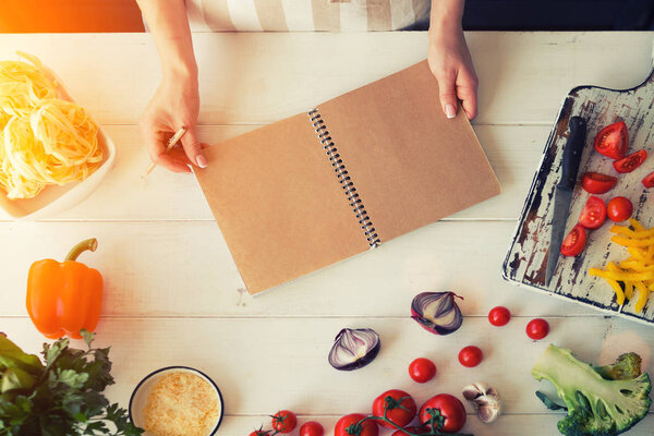 Young woman is holding cookbook and writing a new recipe. Female chef with cookbook. Cooking, culinary, food and people concept. Fresh organic vegetables and spices on a wooden background. Heatly food