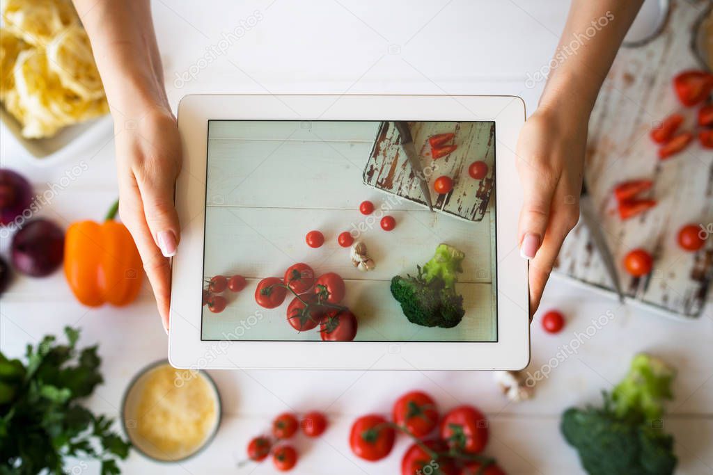Close up view of woman's hand holding modern touch pad with just made picture of kitchen table with colorful vegetables. Cropped view of female blogger photographing cooking process for food recipe.