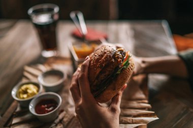 Kadın elleri ve patates kızartması ile burger Cafe arka plan üzerinde tutar. Kraft kağıt Barbekü, taze hamburger pişmiş. Amerikan Mutfagi. Et ve sebze closeup vasıl geçmiş odaklanmamış ile büyük hamburger