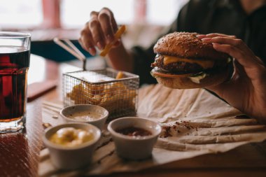 Çizburger tutarak ve serinletici içecek kadın. El lokanta hamburger, Amerikan yemek tutan genç kız. Sığır eti burger ile patates kızartması rustik ahşap arka plan üzerinde elinde. Seçici odak.