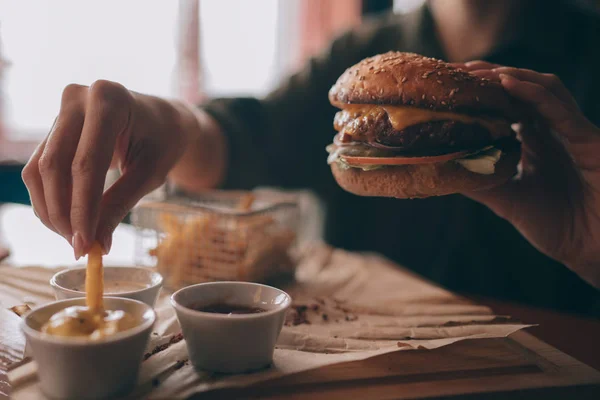 Woman holds burger with hands and fries on the background in cafe. Fresh burger cooked at barbecue in craft paper. American food. Big hamburger with meat and vegetables closeup unfocused at background