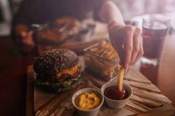 Tasty burger and sauce on wooden tray. Woman eating burger and chips in cafe. People and eating concept. Hamburger, french fries, ketchup, mustard on a wooden board. Toned image.