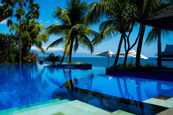 Marble steps leave under pool water. A beach on tropic coast, white beach umbrellas, sunbeds and palm trees against the sea. Natural garden in luxurious resort. Flower trees and green plants.