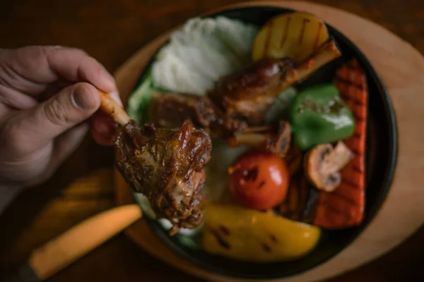 Eating and leisure concept. Dinner on the table. Tasty grilled lamb leg with vegetables and herbs on a roasting iron pan. Assorted delicious vegetables with grilled meat. Toned image. Selective focus.