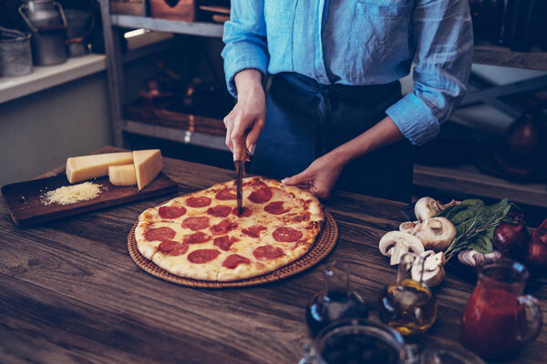 Woman cutting fresh baked homemade pizza on rustic kitchen background. Cut into slices delicious pizza with mushrooms and ham. Cheese and tomatoes on wooden table. Healthy foods, cooking concept.
