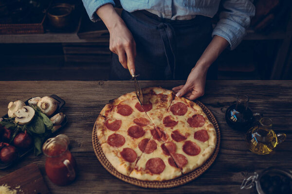 Woman cutting fresh baked homemade pizza on rustic kitchen background. Cut into slices delicious pizza with mushrooms and ham. Cheese and tomatoes on wooden table. Healthy foods, cooking concept.