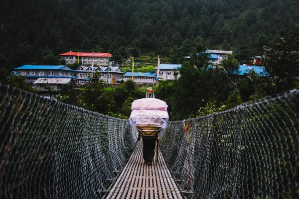 Nepalese porter sherpa carrying heavy sacks and crossing a suspension bridge in Everest Region. Sagarmatha national park, Himalaya, Nepal. Sherpas are elite mountaineers and experts in the mountains. — Stock Image Nepalese Porter Sherpa Carrying Heavy Sacks Crossing Suspension Bridge Everest Stock Photo