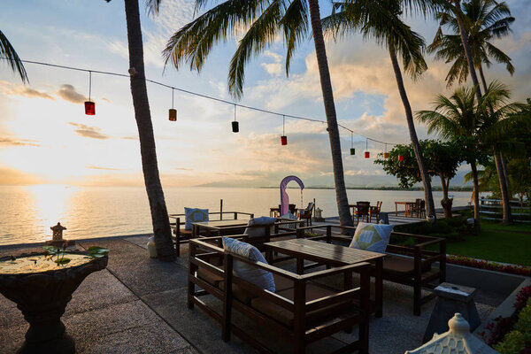 Sunset over coastline. Romantic sunset on the shore of a tropical island. Restaurant on the beach. Empty  table and chairs under palm trees at restaurant on open terrace. Ocean lagoon on background.