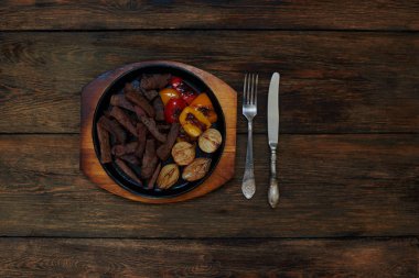 Hangi cool taze hazırlanmış kuzu eti ve bir ızgara, kızarmış sebze yalan yanında gümüş çatal bıçak takımı bir düz dökme demir tava üzerinde karanlık bir ahşap tablodur